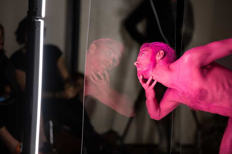 Dancer Jason Collins juts out his chin, taking it in his hand. He stares out toward the audience through a clear plastic wall.